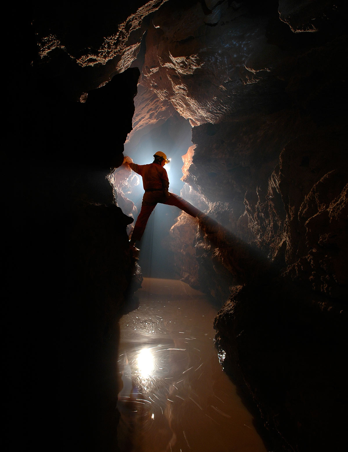 Caver straddling an underground stream in a narrow cave passage, backlighting for effect.